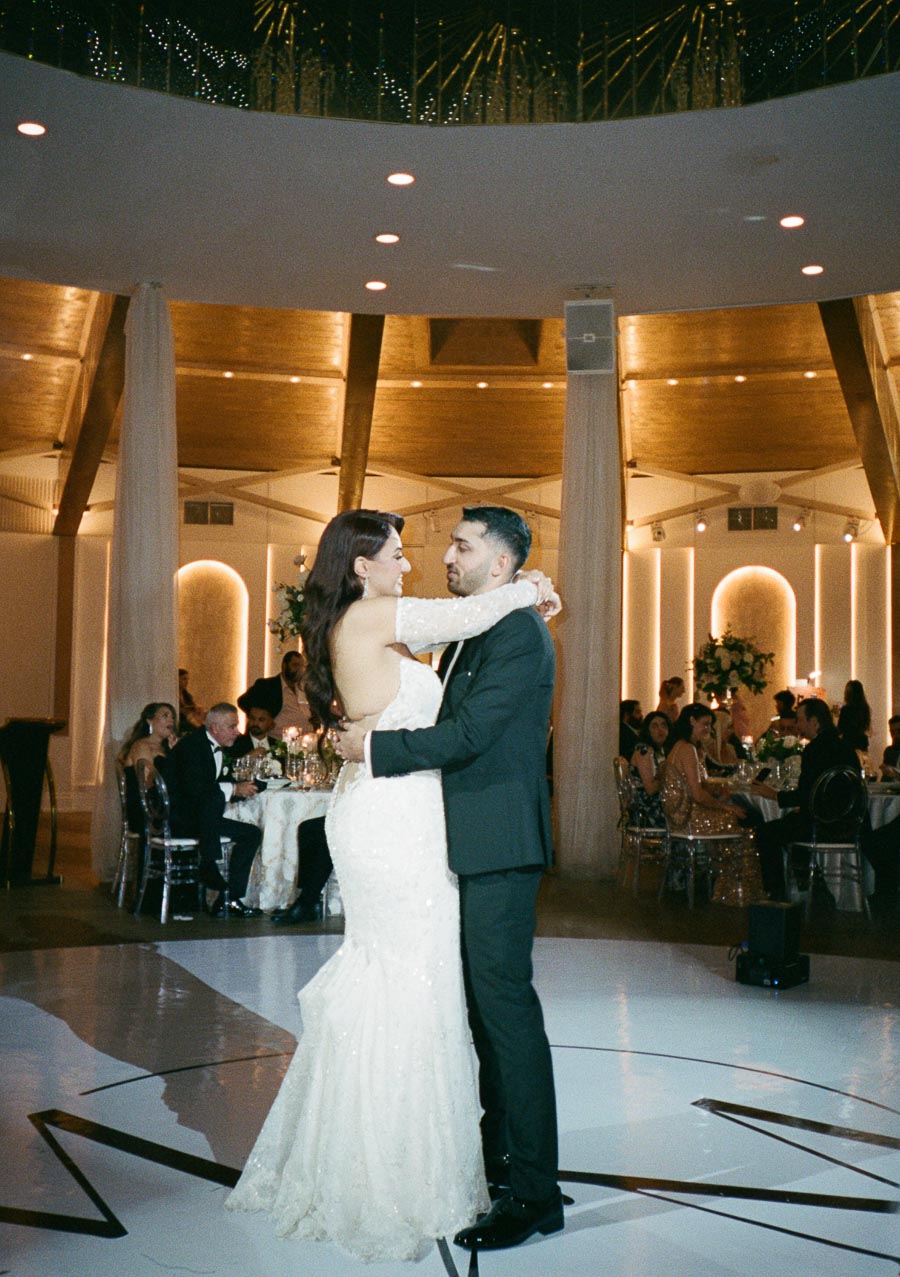A bride and groom share their first dance at a beautifully decorated wedding reception, surrounded by elegantly dressed guests seated at candlelit tables in a grand venue with a high ceiling and warm lighting.