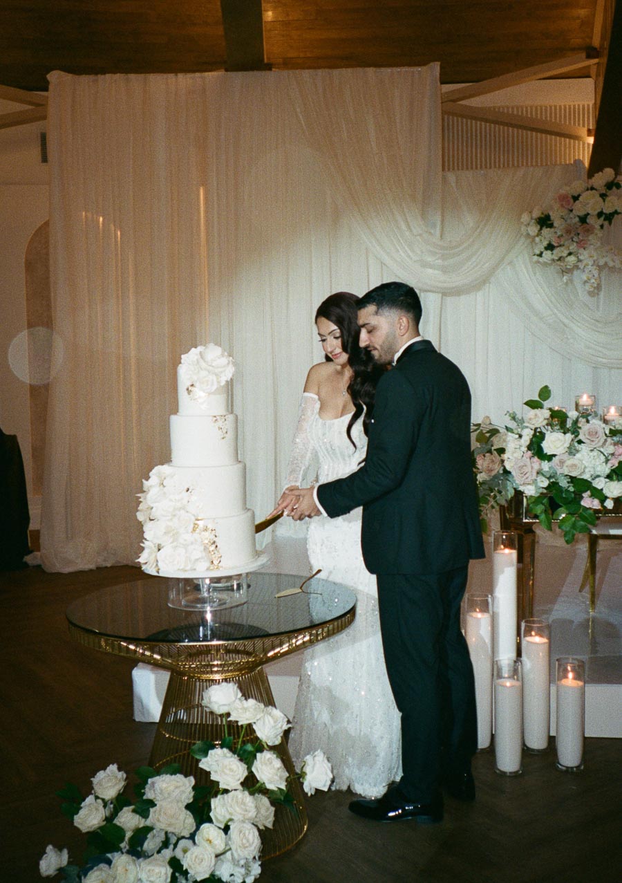 Bride and groom cutting a three-tiered white wedding cake adorned with floral decorations, surrounded by candles and flower arrangements in an elegant reception setting.