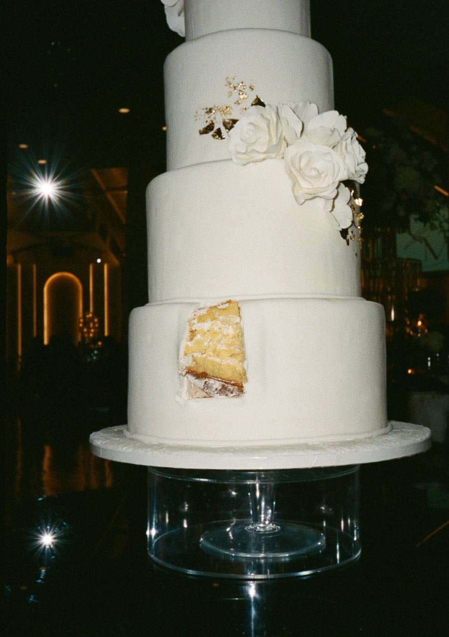 Four-tier white wedding cake with a slice removed, decorated with white roses and gold accents, displayed on a clear stand in a dimly lit venue.