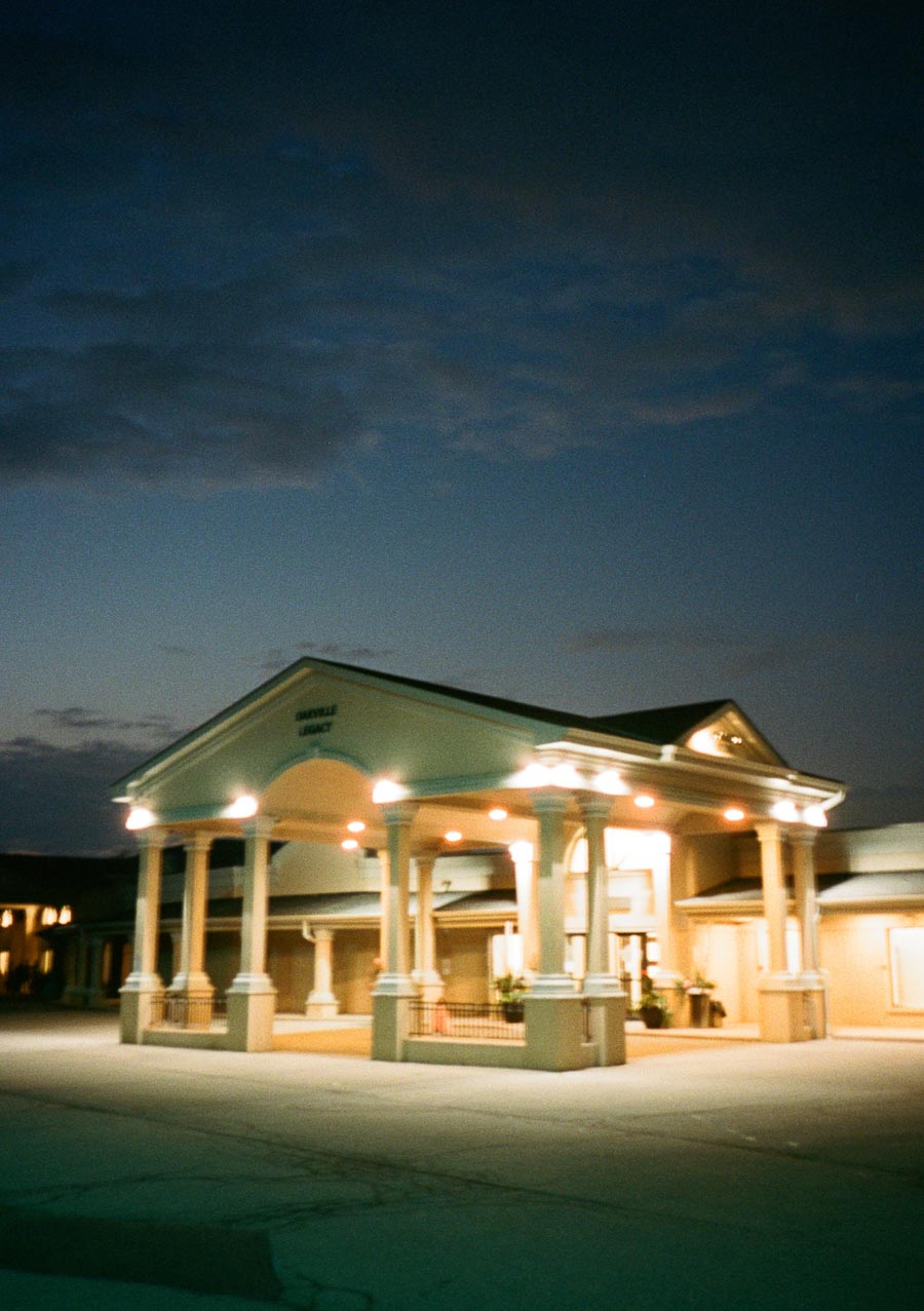 Illuminated building entrance at dusk with white pillars and a triangular roof, set against a deep blue sky.