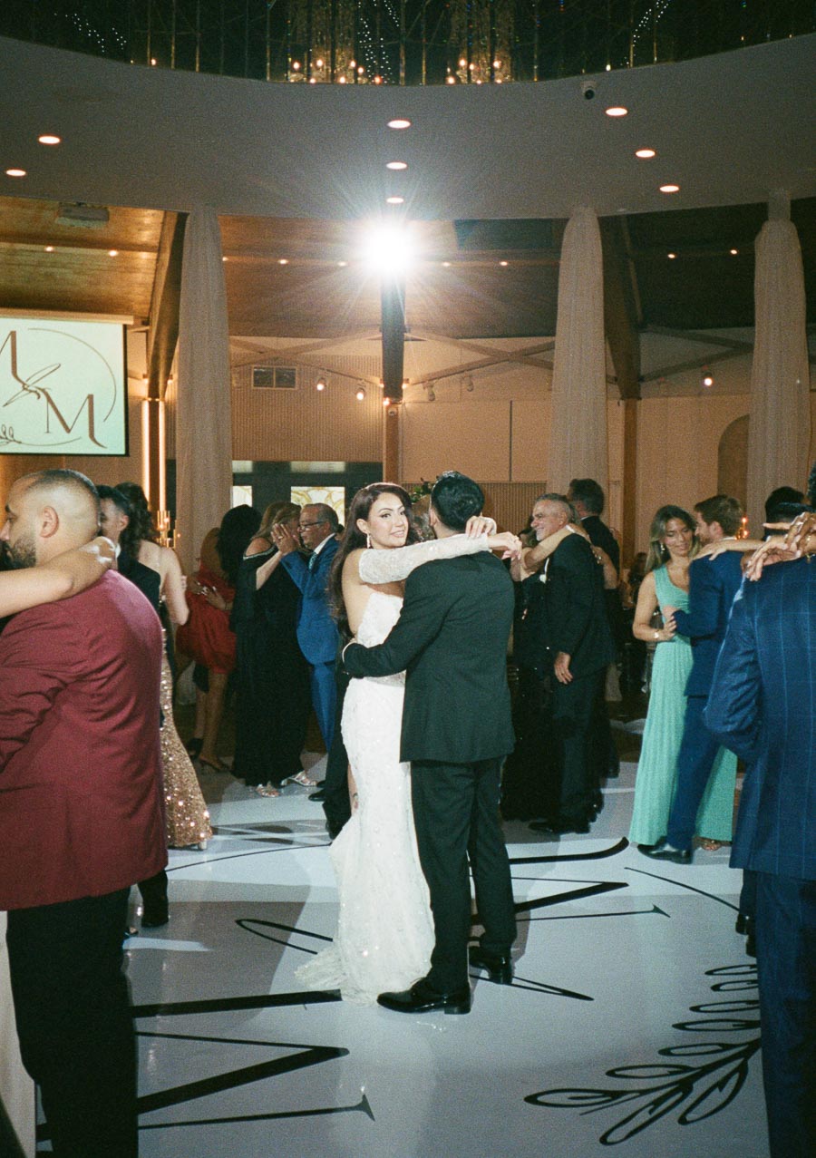 A joyful couple dances at their wedding reception surrounded by elegantly dressed guests, under a softly lit and stylish venue ceiling.