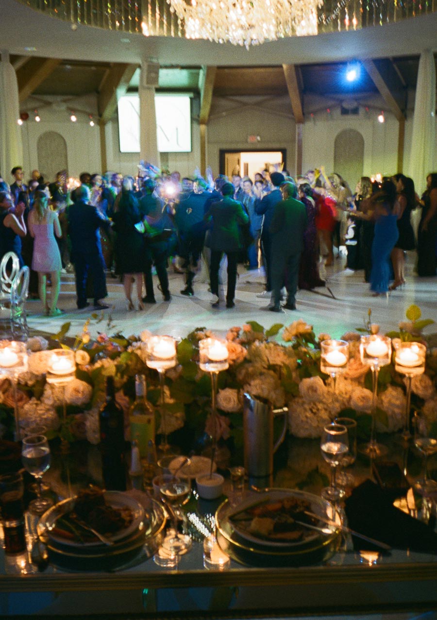 A lively wedding reception scene with a large group of people dancing under a bright chandelier. The foreground shows a beautifully set dining table adorned with elegant floral arrangements and candleholders, creating a warm and festive atmosphere.