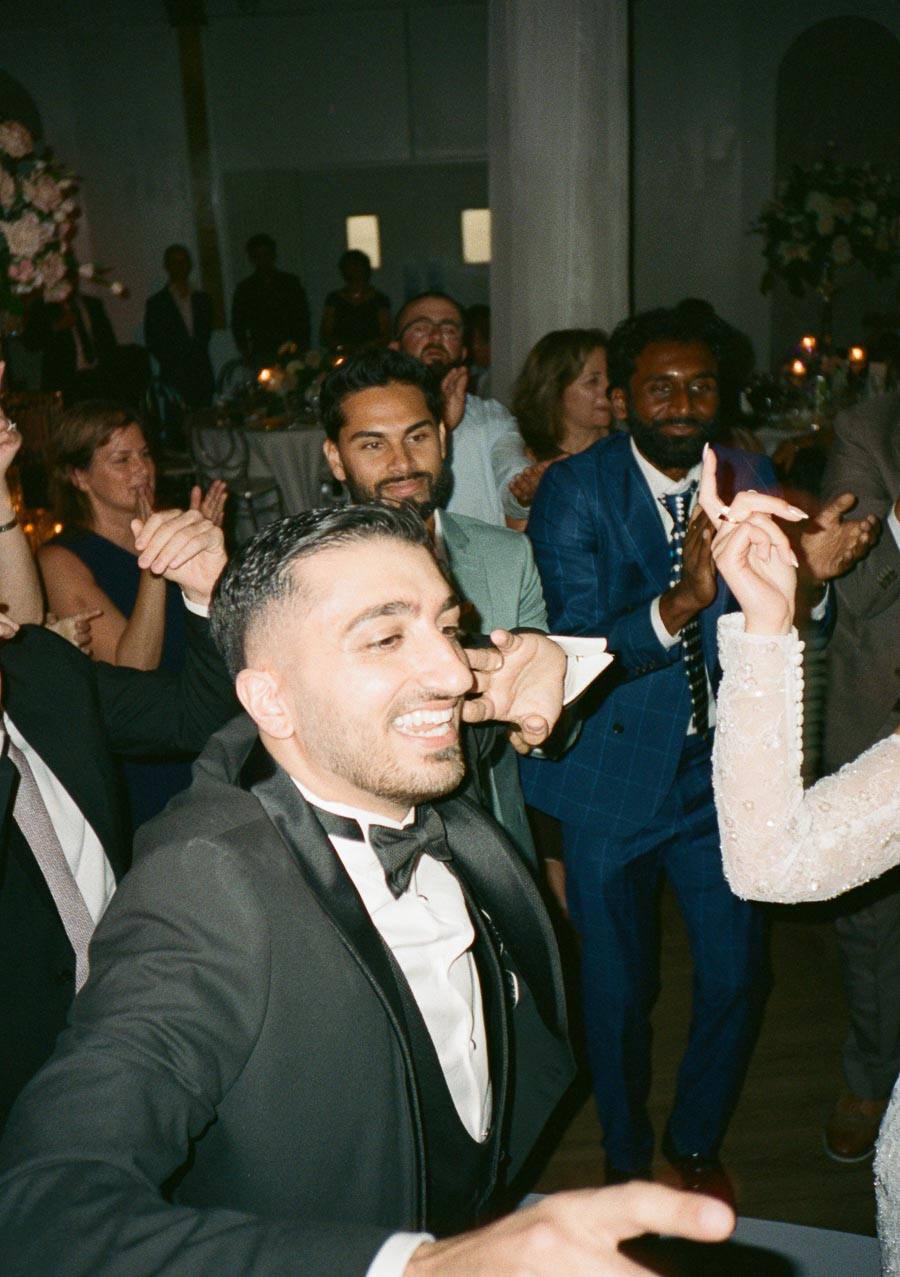 A joyful wedding celebration with a group of well-dressed guests dancing energetically at a reception, featuring elegant attire and a lively atmosphere.