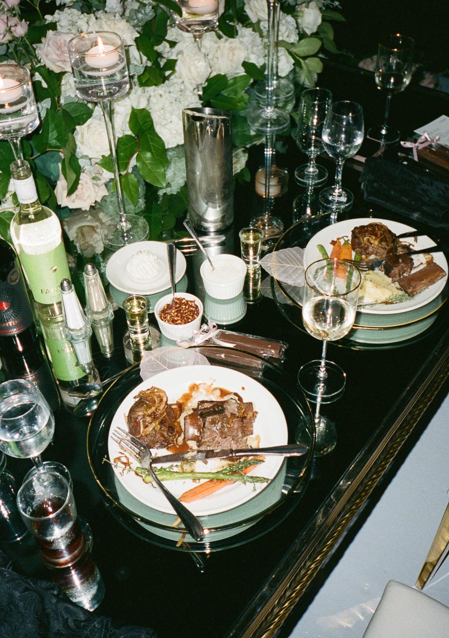 Elegant dinner table setup with gourmet dishes of beef, vegetables, and mashed potatoes, surrounded by wine bottles, candles, and floral centerpieces.