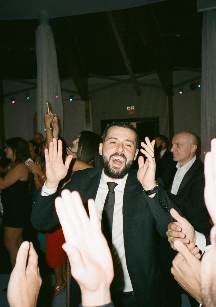 A bearded man in a suit smiling and dancing at a lively indoor event, surrounded by a crowd clapping and celebrating.