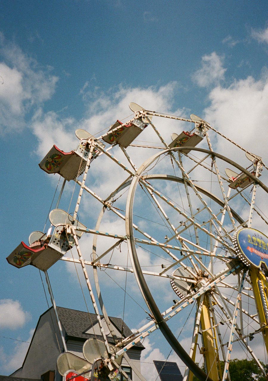 Vintage Ferris wheel against a blue sky with fluffy clouds, featuring colorful passenger seats at a carnival, with a building and trees visible in the background.