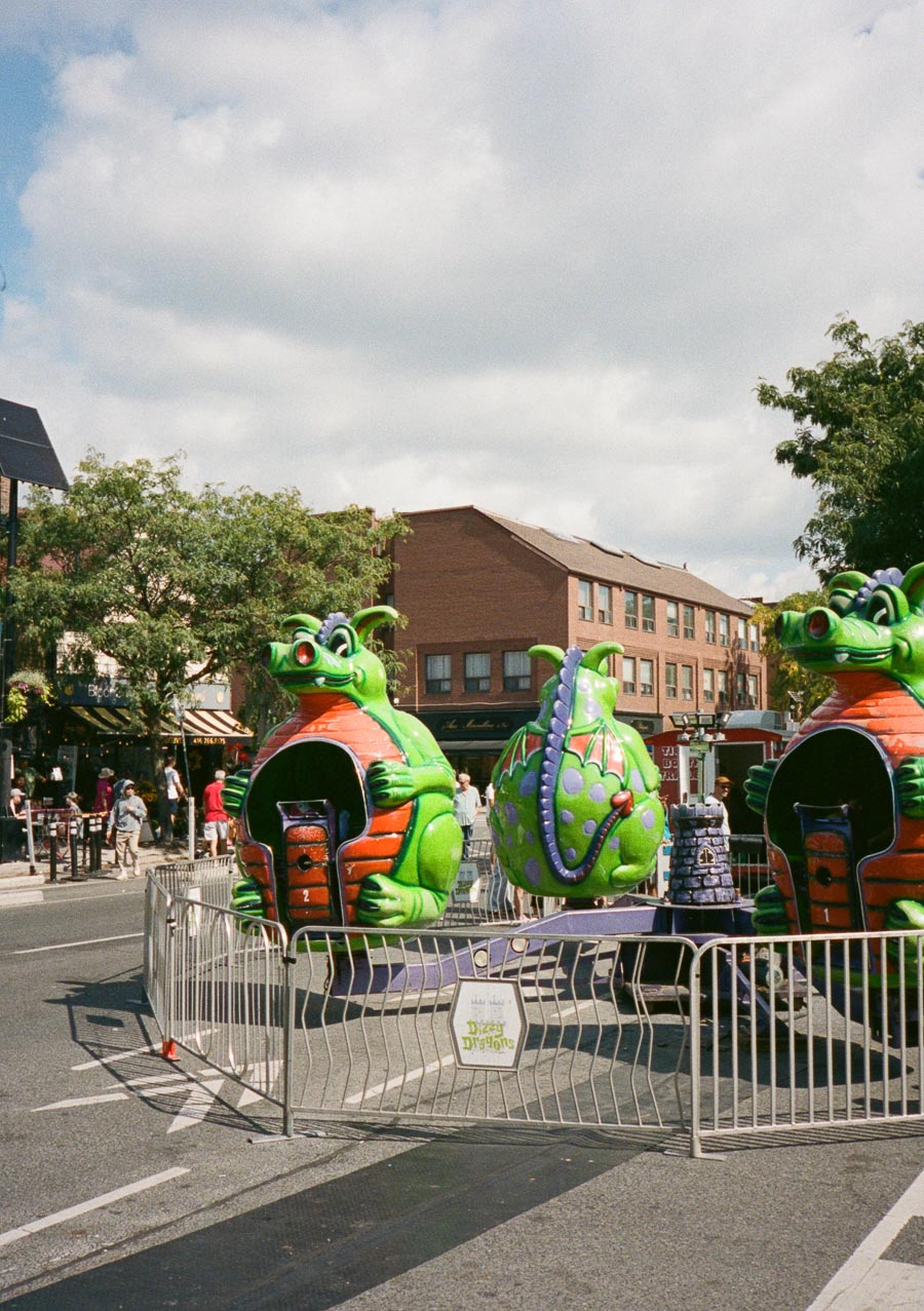 Inflatable dragon-themed ride at a street fair, surrounded by a metal fence, with a brown brick building and trees in the background under a partly cloudy sky.