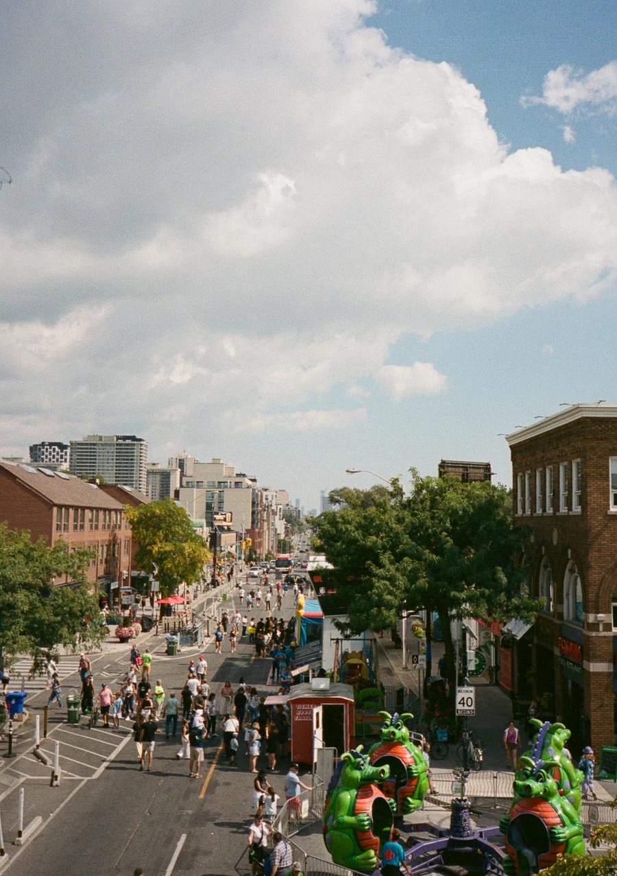 A bustling urban street during a daytime festival, with people enjoying activities and carnival rides, surrounded by buildings and lush green trees, under a partly cloudy sky.
