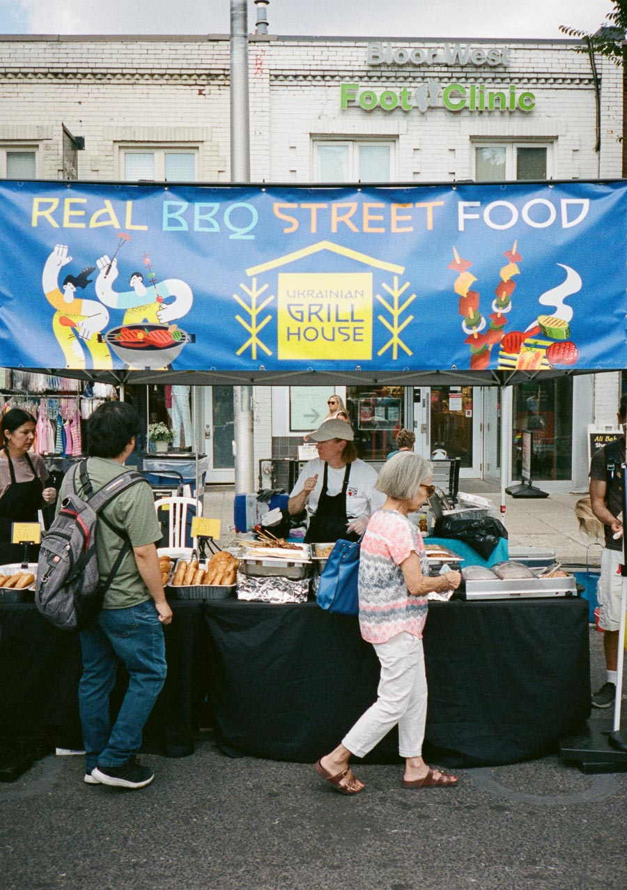 A vibrant street food market featuring a Ukrainian BBQ stand with a colorful banner reading Real BBQ Street Food and Ukrainian Grill House. Customers browse freshly prepared food displayed on tables under a blue canopy, enhancing the lively atmosphere of the outdoor market.
