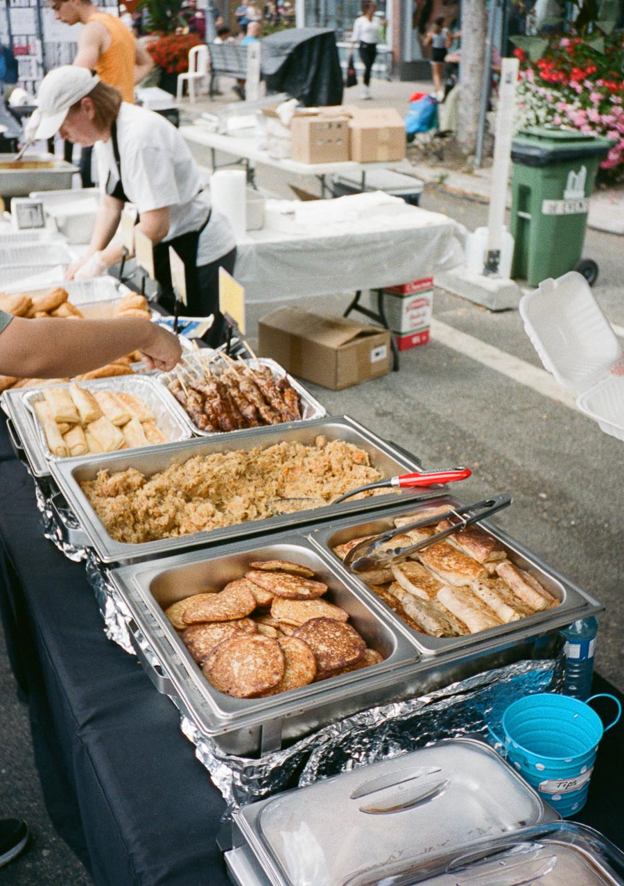 Street food market stall featuring a variety of dishes, including rice, cookies, and skewers, with a vendor and customers in the background.