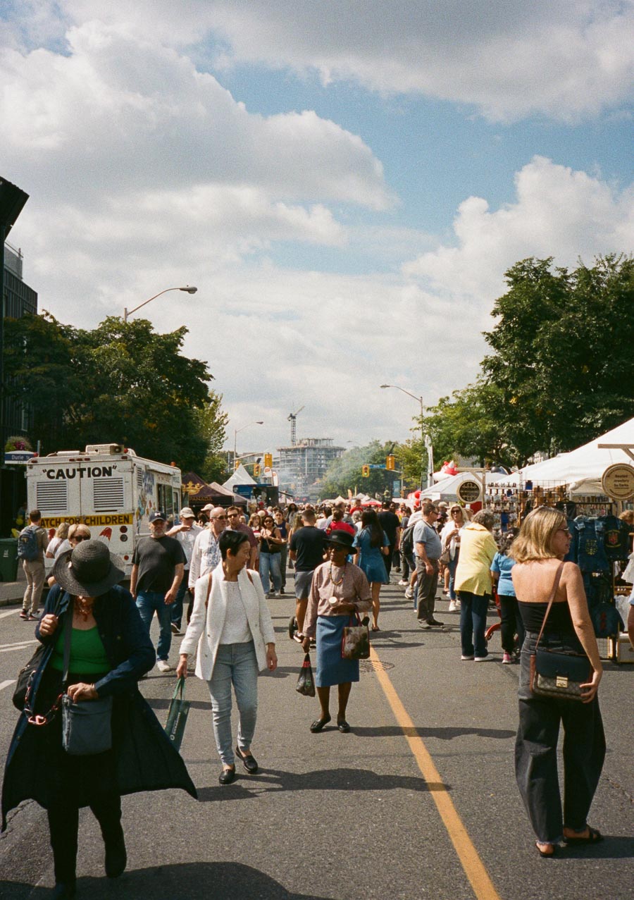 Outdoor street festival with a diverse crowd. People are browsing vendor stalls on a sunny day, featuring food trucks, market tents, and lush greenery under a partly cloudy sky.