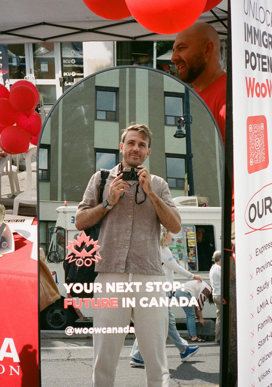 Man taking a selfie in the reflection of a mirror at an outdoor event, surrounded by promotional materials for immigration services in Canada, with red balloons and informational banners visible.