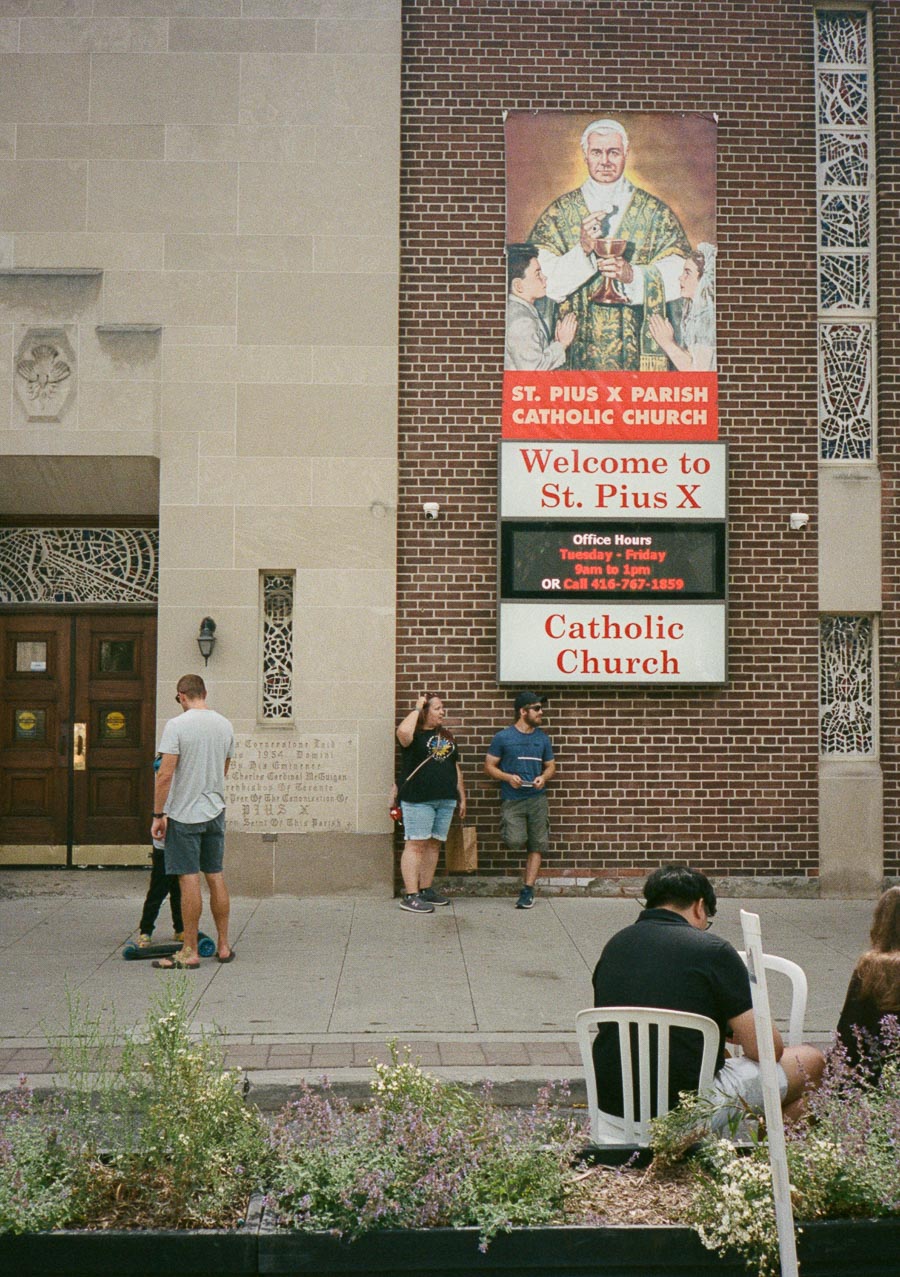 Exterior view of St. Pius X Parish Catholic Church, featuring a prominent welcome sign and a decorative wall with stained glass accents. People are casually standing and sitting outside the entrance, creating a sense of community engagement.