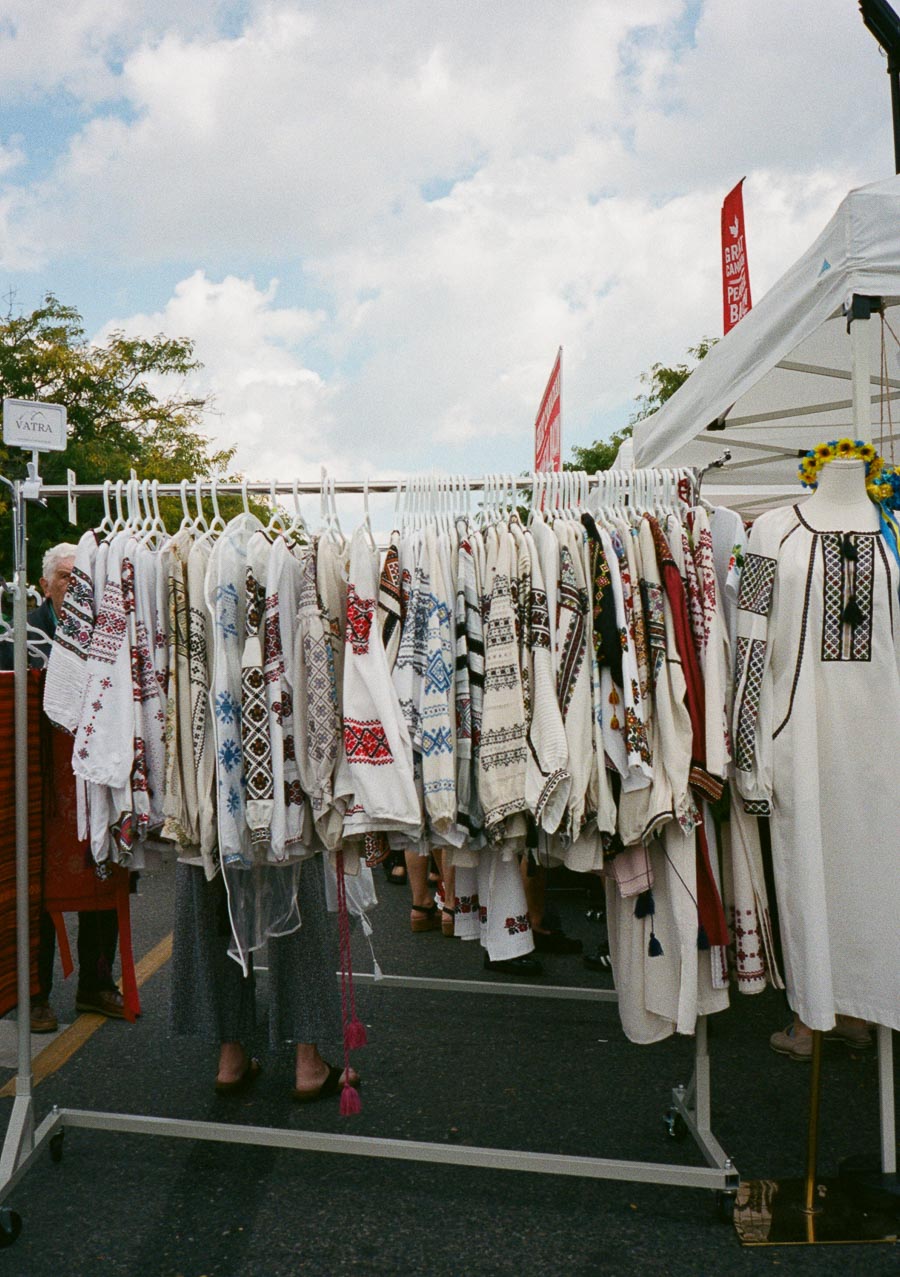 Rack of traditional embroidered blouses at an outdoor market stall under a blue sky, showcasing Eastern European folk clothing patterns in vibrant colors.