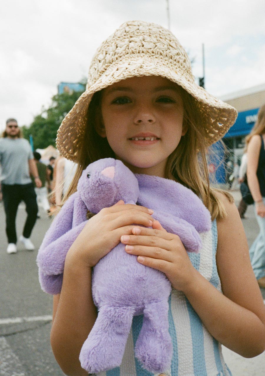 Young girl wearing a straw hat, holding a purple stuffed animal outdoors on a sunny day.