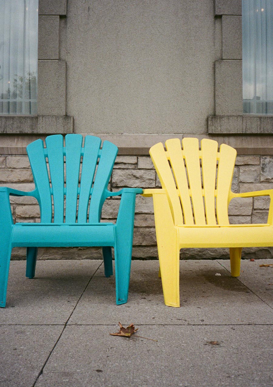 Two vibrant Adirondack chairs in blue and yellow sit on a concrete sidewalk against a stone building exterior, capturing a cozy outdoor setting.