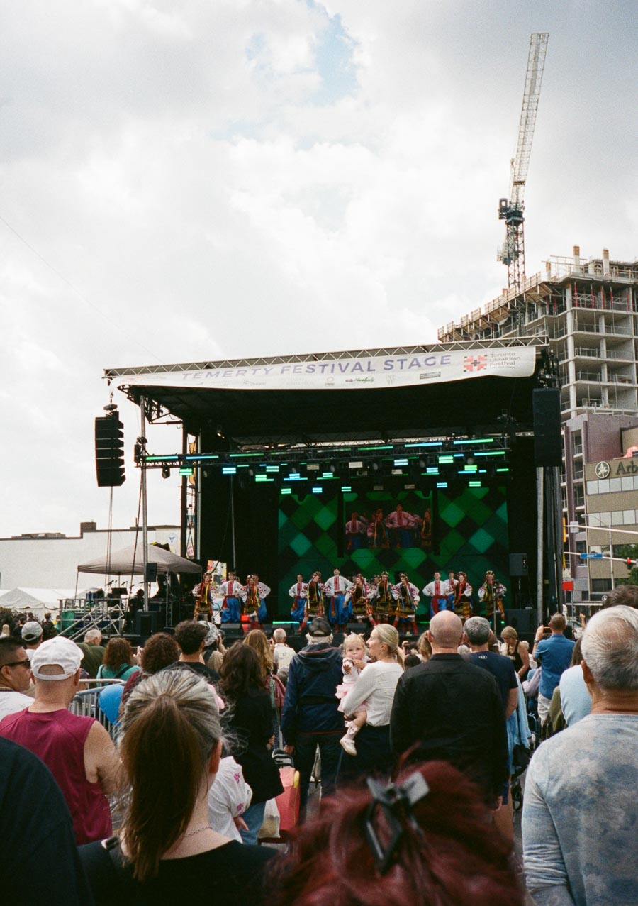Outdoor festival performance on the Temerty Festival Stage featuring a group of dancers in colorful traditional costumes, with a large crowd gathered under a partly cloudy sky in an urban setting.