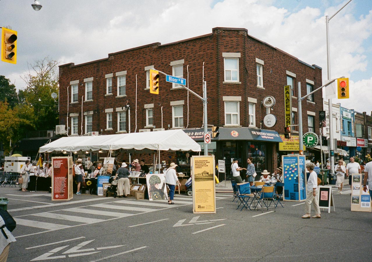 Street festival at a bustling city intersection with people exploring vendor stalls under a white canopy. A brick building with various storefronts, including a Lebanese food shop and a burger place, serves as the backdrop. Pedestrians walk across the crosswalk, and informational displays are set up throughout the area. Street signs and traffic lights are visible.