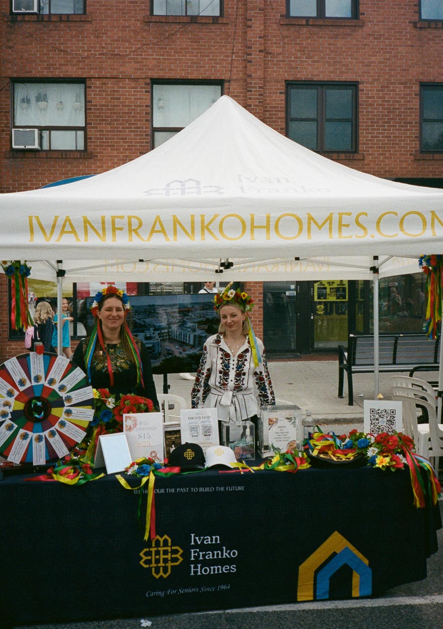 Outdoor stall representing Ivan Franko Homes at a community event with two people in traditional attire, promotional materials, and colorful decorations, set against a brick building background.