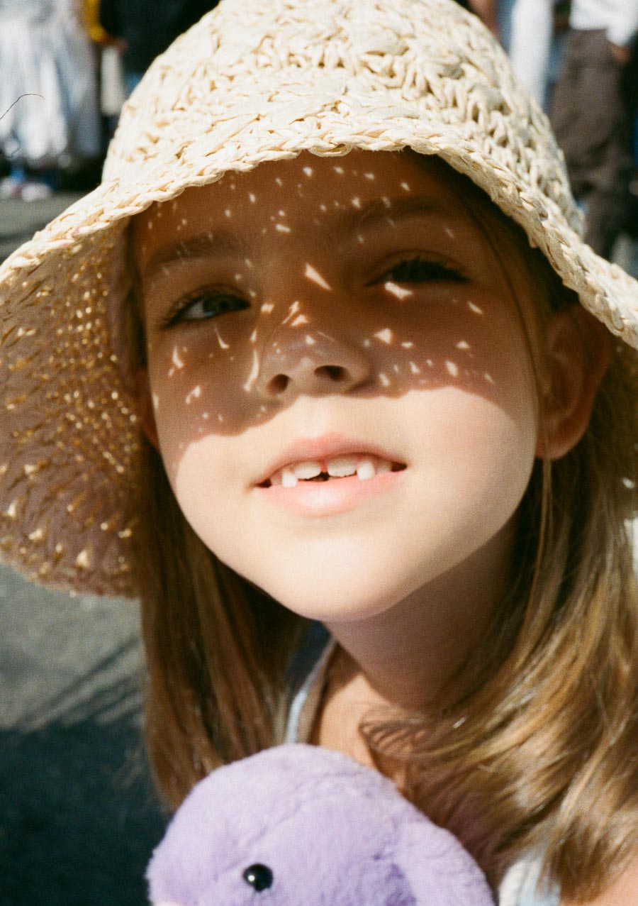 A young girl wearing a straw hat smiles warmly while holding a purple plush toy, with sunlight creating patterns through the hat's weave.