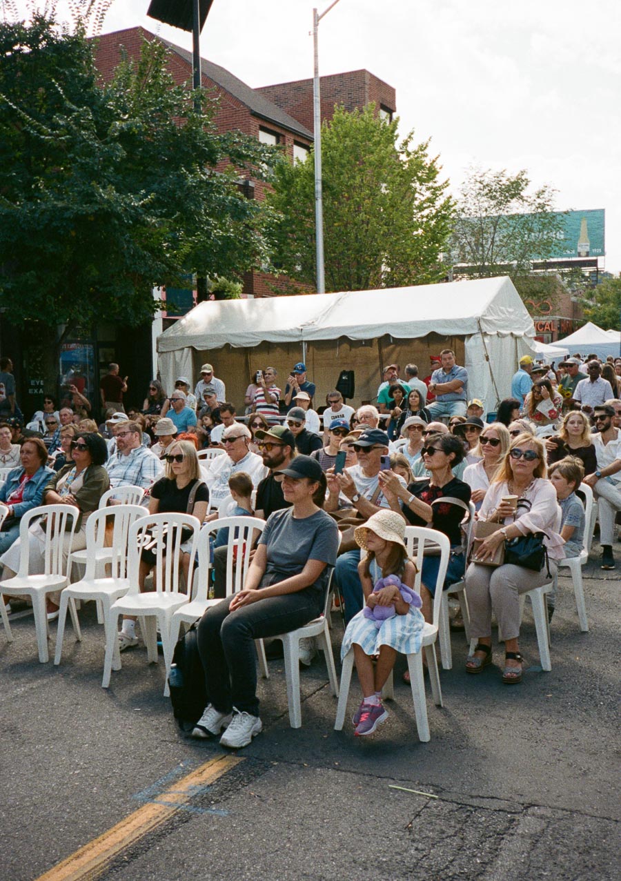 A diverse group of people sits together on white chairs under a sunny sky at an outdoor festival. They are positioned near a large white canopy tent, surrounded by trees and brick buildings, suggesting a lively community event in an urban setting.