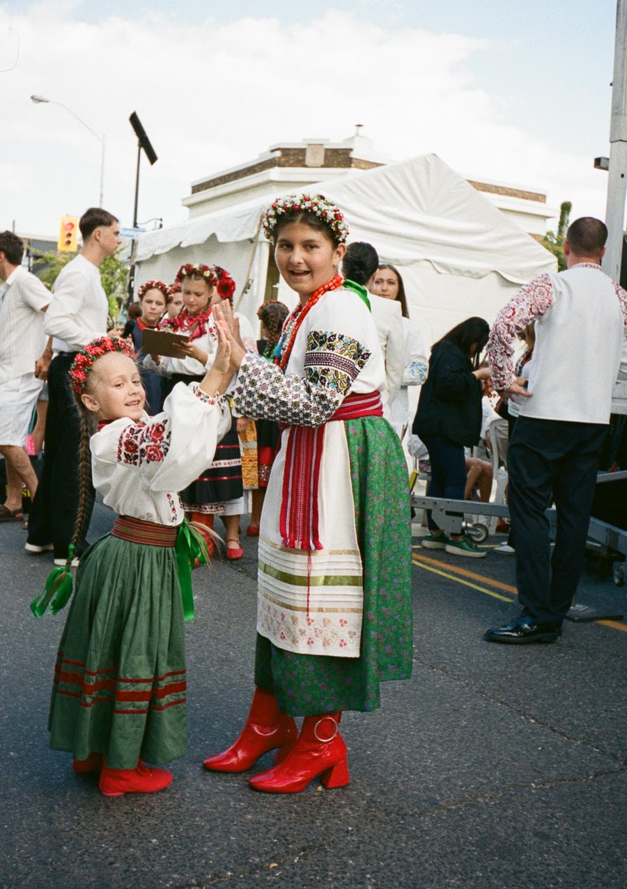 Two young girls in traditional Ukrainian attire perform a folk dance at an outdoor cultural festival, surrounded by people in traditional costumes.