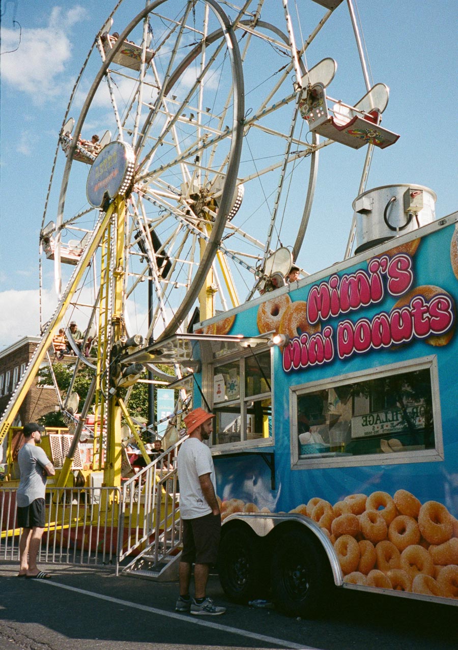 Ferris wheel next to Mimi's Mini Donuts food truck at a street fair, with sunny sky and people enjoying the attractions.