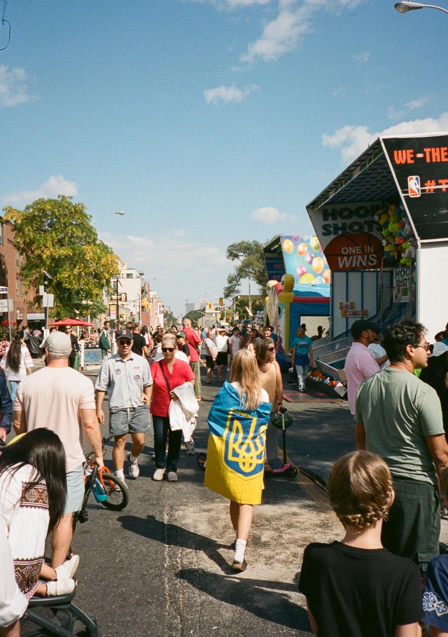 Street fair scene with a diverse crowd on a sunny day, featuring carnival games and a person wearing a blue and yellow flag.