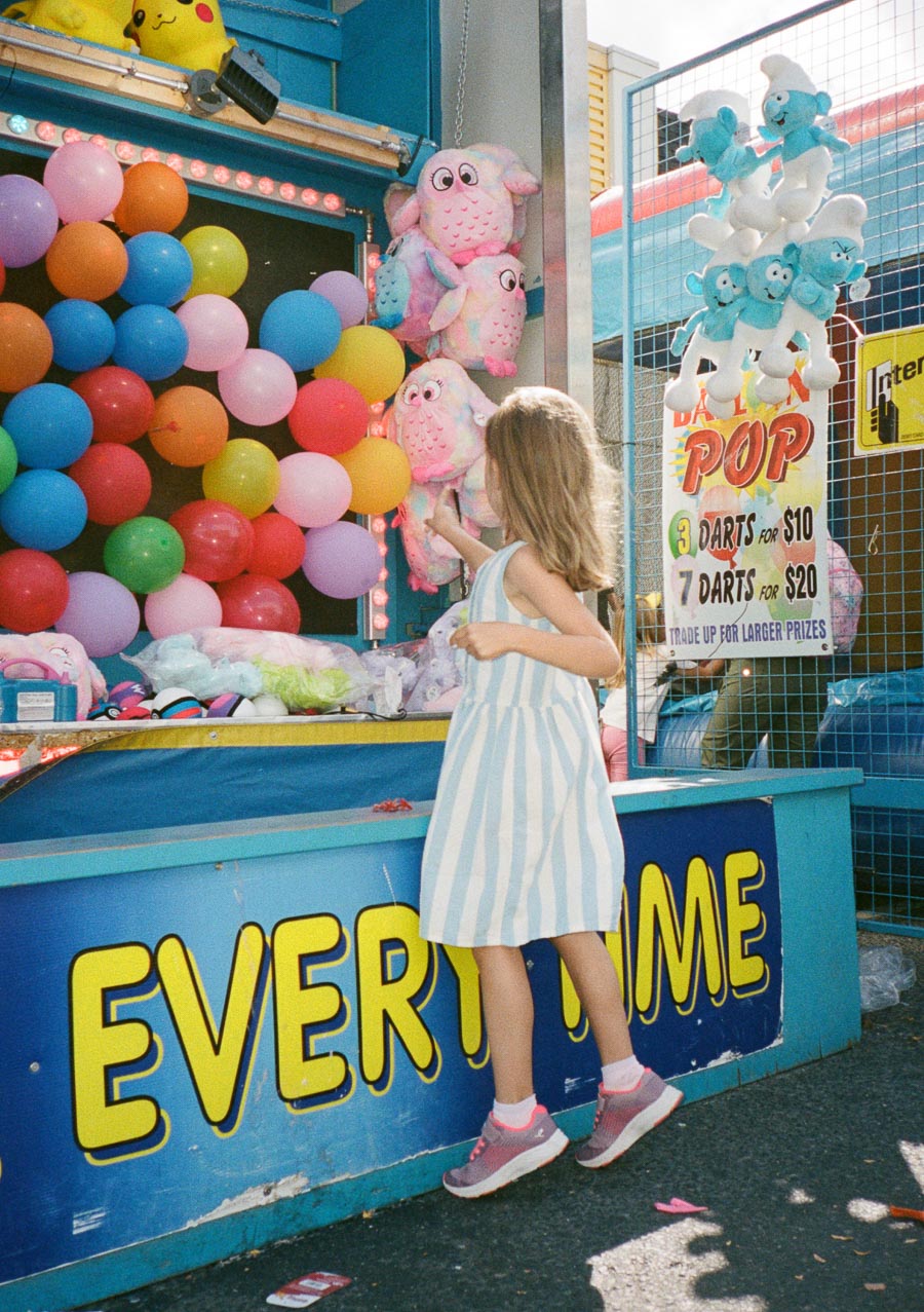 Young girl in a striped dress playing a colorful balloon dart game at a carnival, surrounded by plush toys and a price sign, under a sunny sky.