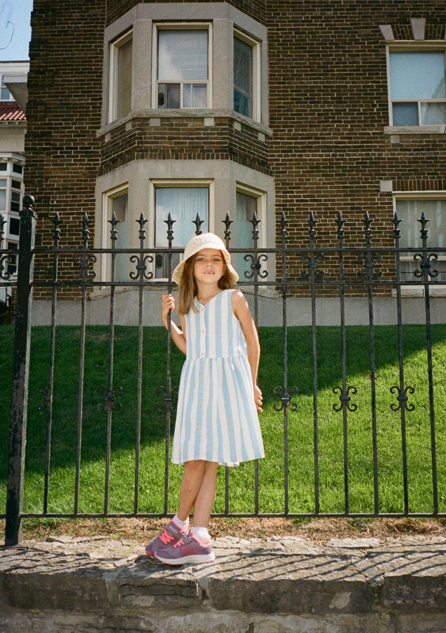 A young girl in a blue and white striped dress stands in front of a wrought iron fence, wearing a sun hat and pink sneakers, with a brick building and green lawn in the background.