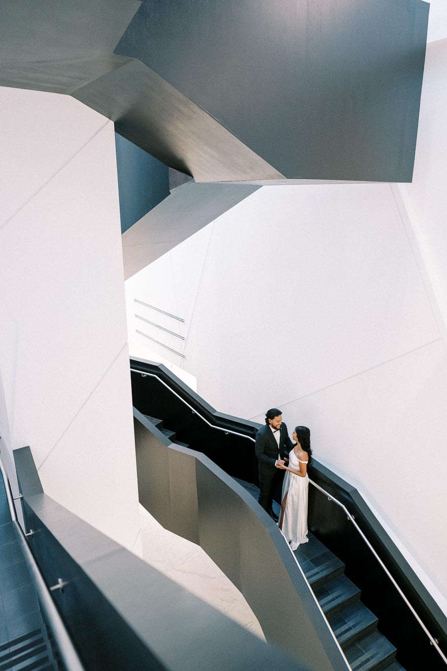 A couple in formal attire standing on a modern black staircase against a minimalist architectural background with clean white walls and geometric angles.