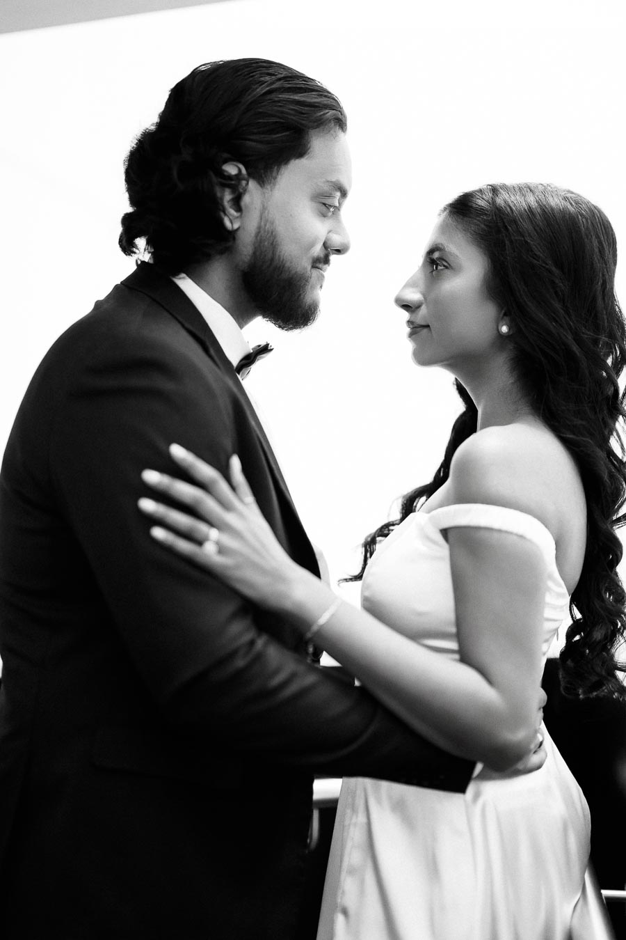 A black and white image of a couple elegantly dressed for a formal event, with the woman in a strapless gown and the man in a suit and bow tie, gazing into each other's eyes, conveying a sense of romance and connection.