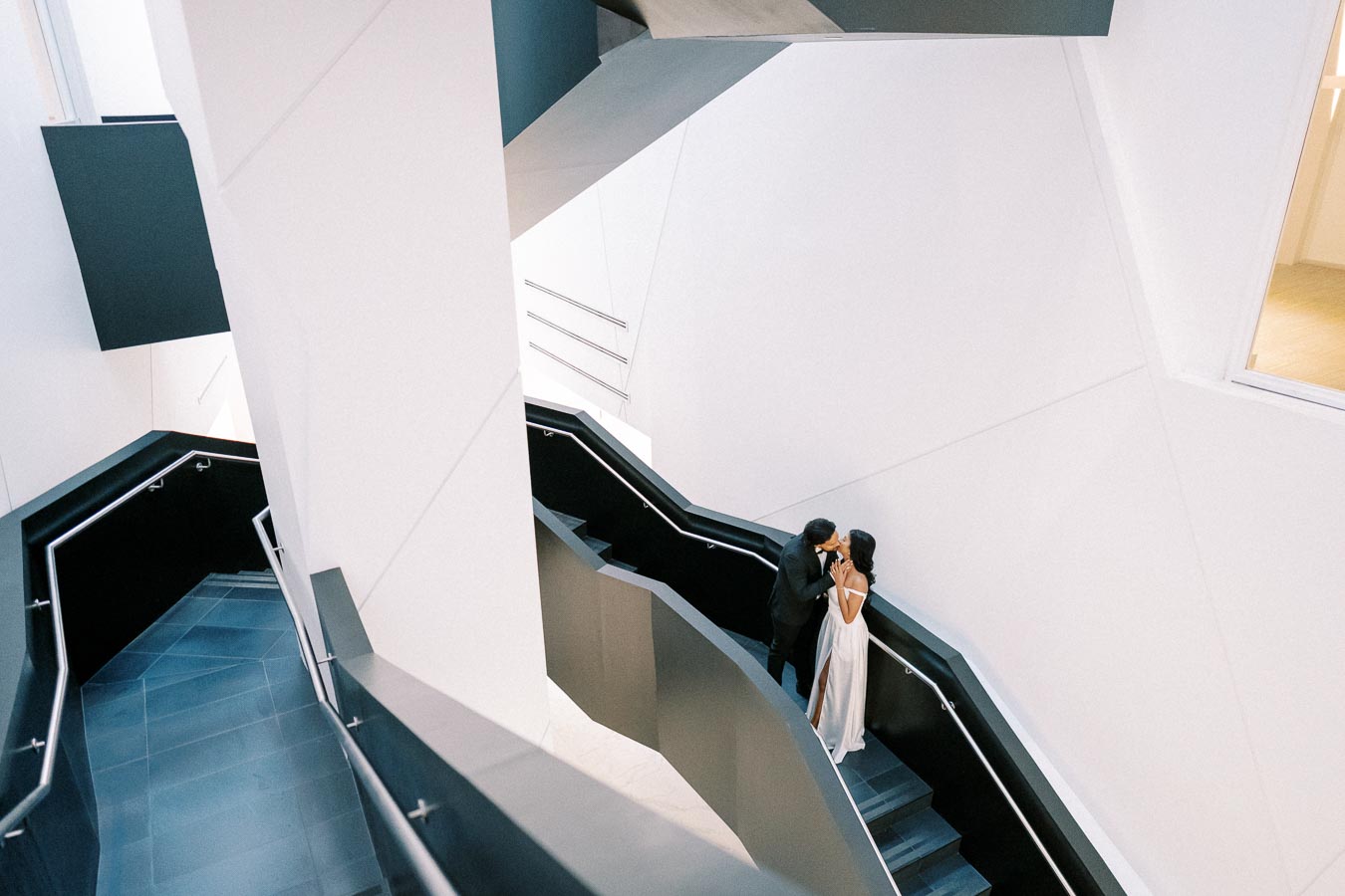 A bride and groom share a romantic moment on a modern staircase with abstract architecture, featuring clean lines and minimalistic design.