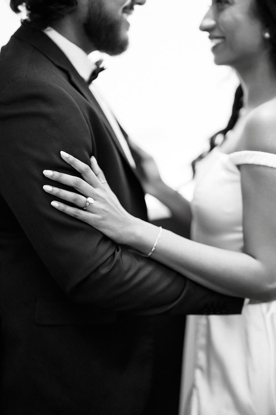 A black and white photo of a couple embracing, with a focus on the woman's elegant engagement ring and wedding dress as they share a joyful moment.