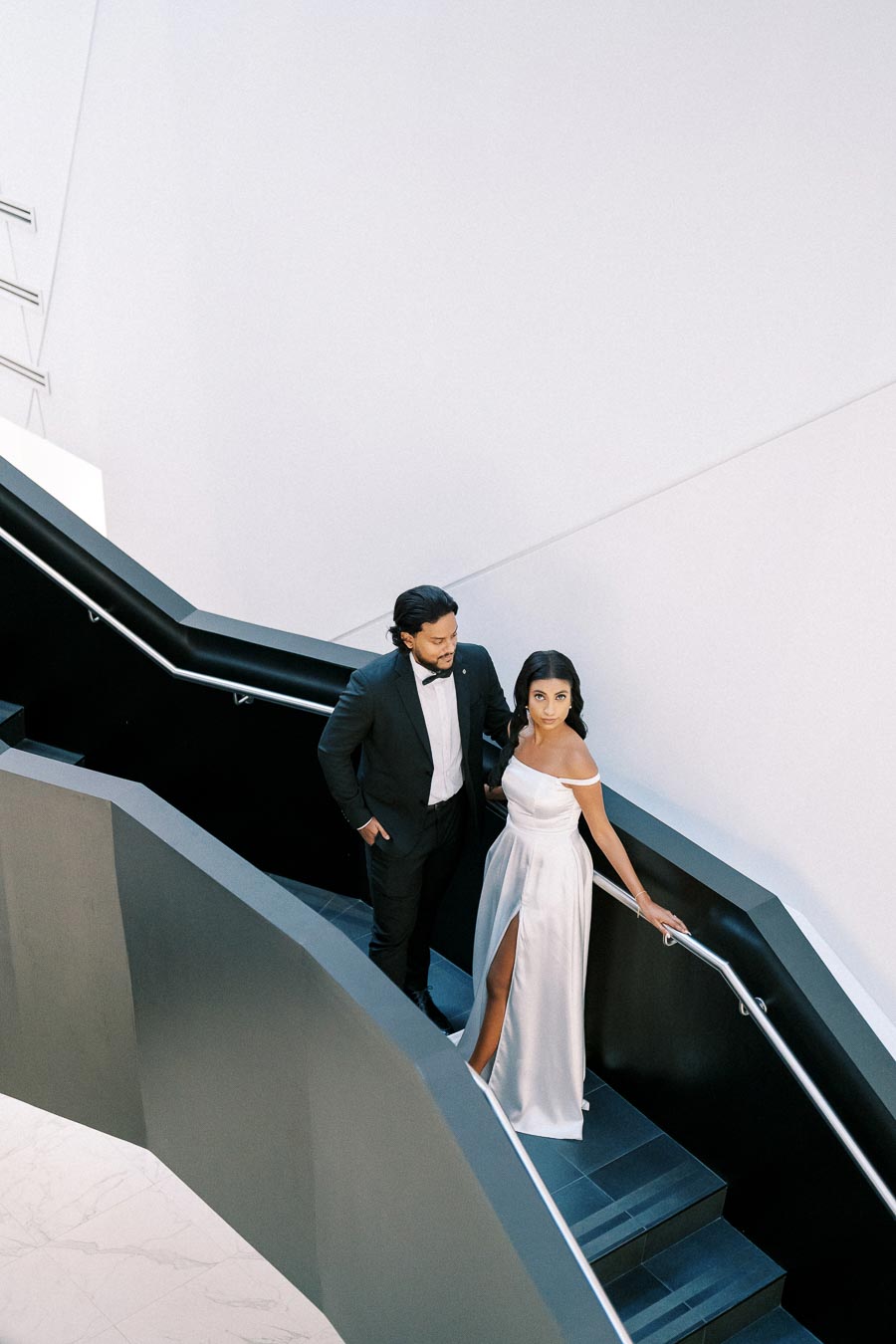 Elegant couple ascending a modern staircase, woman in an off-the-shoulder white gown and man in a tuxedo, showcasing sophisticated evening fashion.