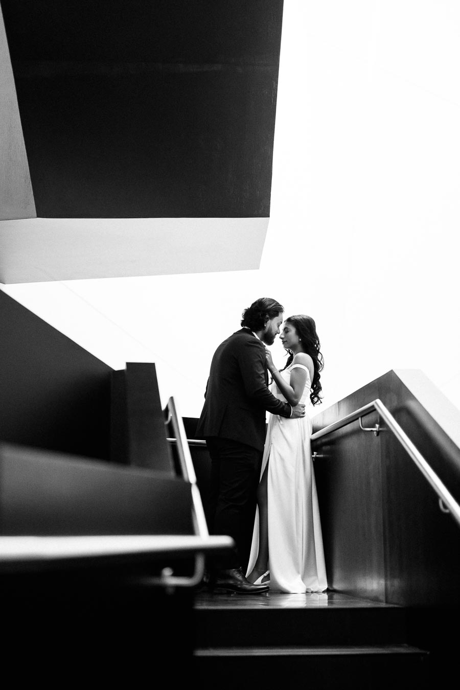 Black and white photo of a couple embracing on a modern staircase, capturing a romantic and intimate moment against an architectural backdrop.