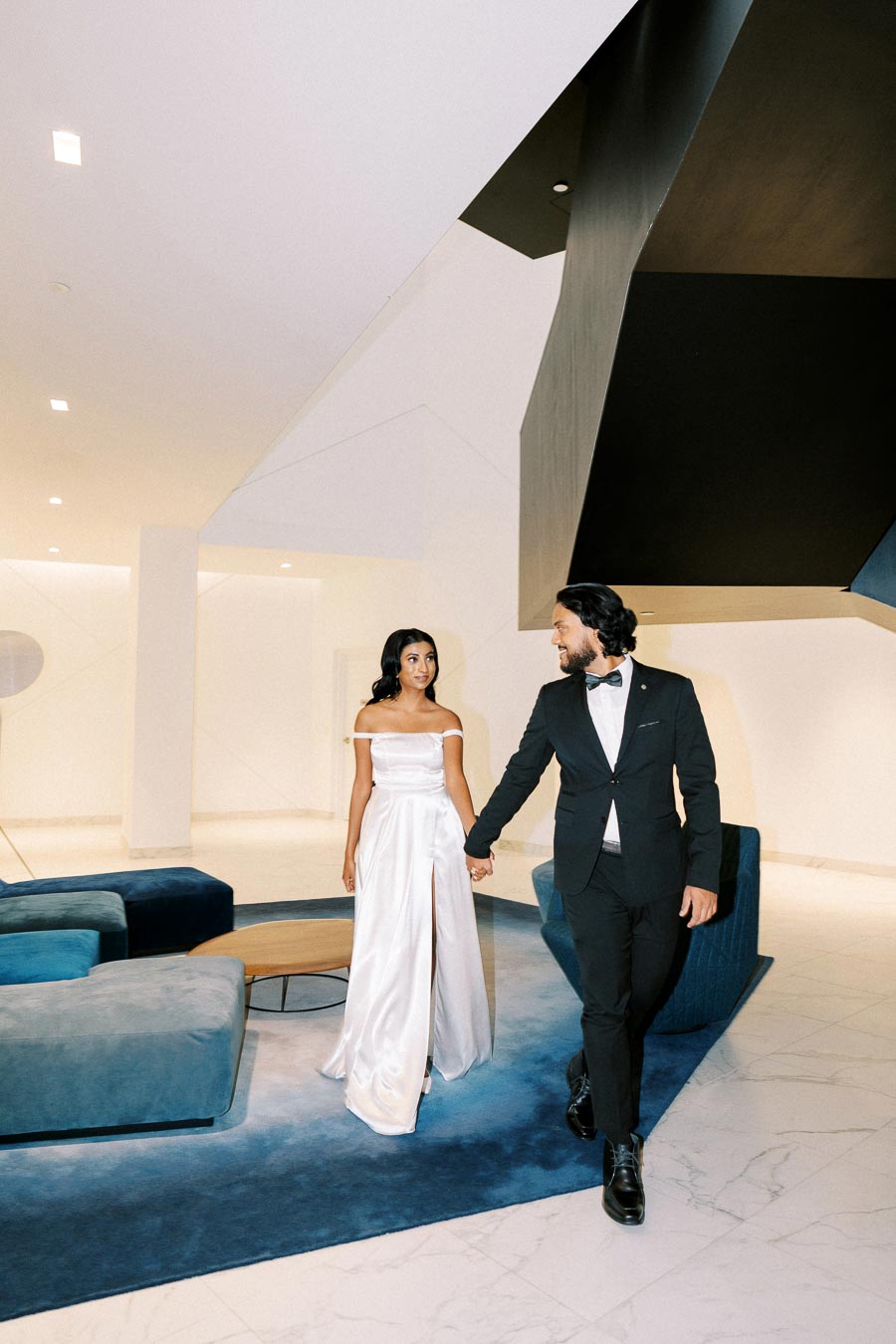 Elegant couple in formal attire walking hand in hand in a modern, minimalist interior with geometric architecture and blue furnishings.