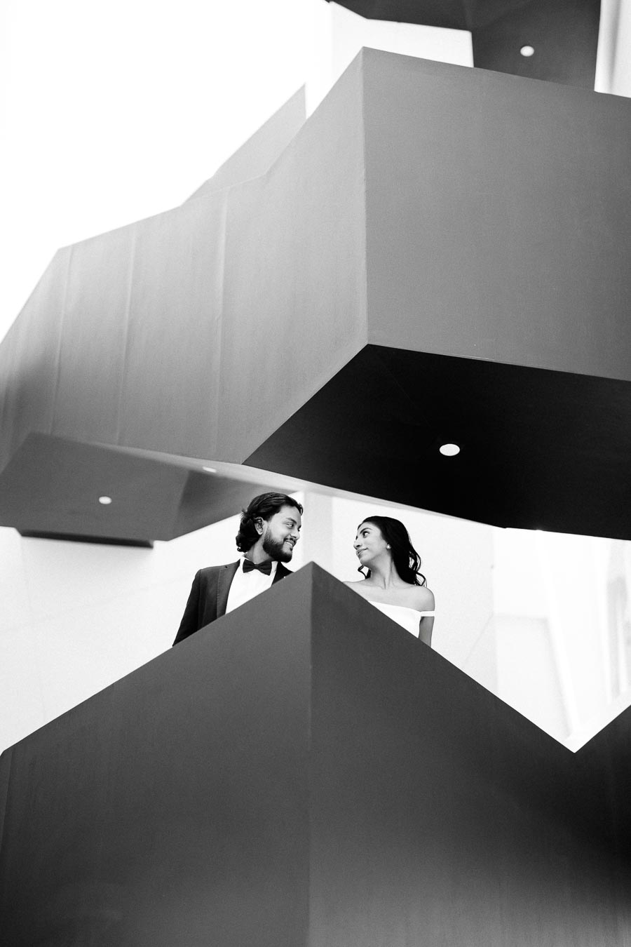 A black and white photo of a couple standing on a modern architectural staircase, gazing at each other and smiling, with geometric shapes and clean lines in the background.