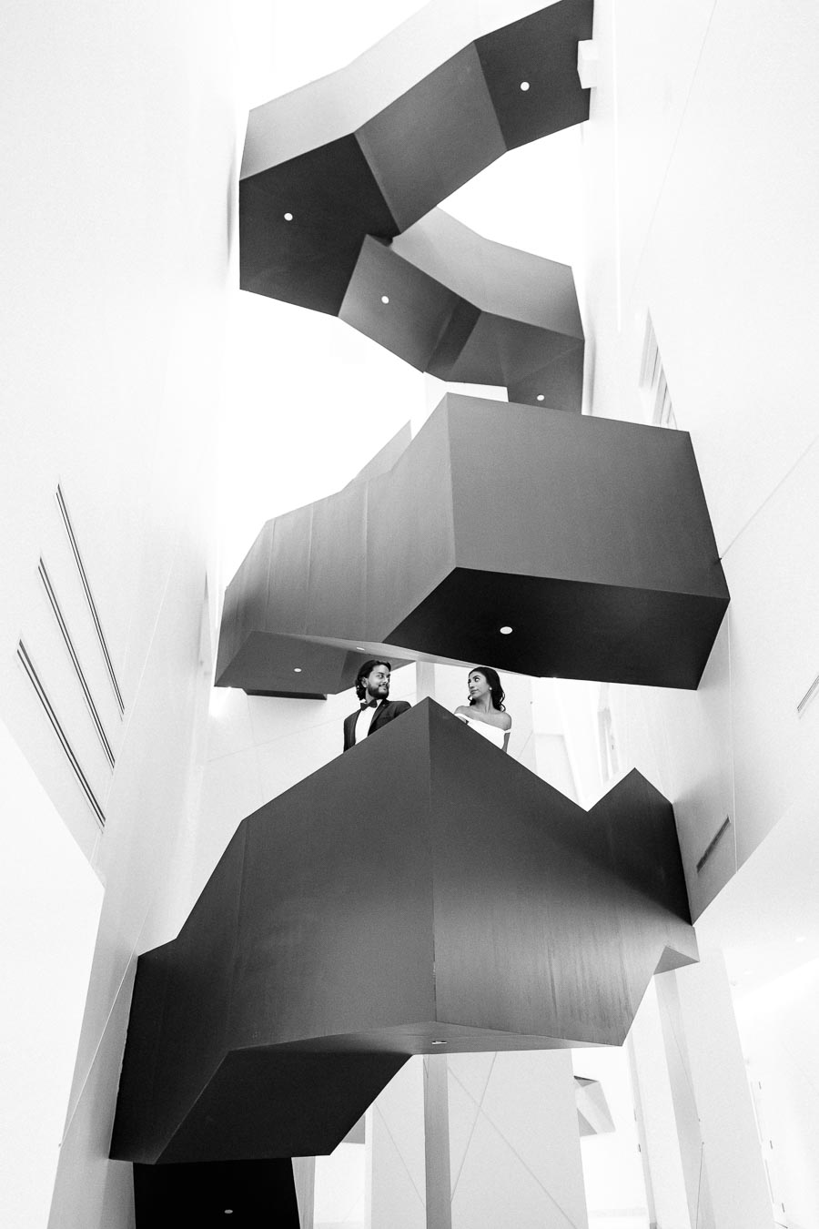 Black and white photo of a modern, geometric staircase with two people standing on a landing, showcasing architectural design and urban elegance.