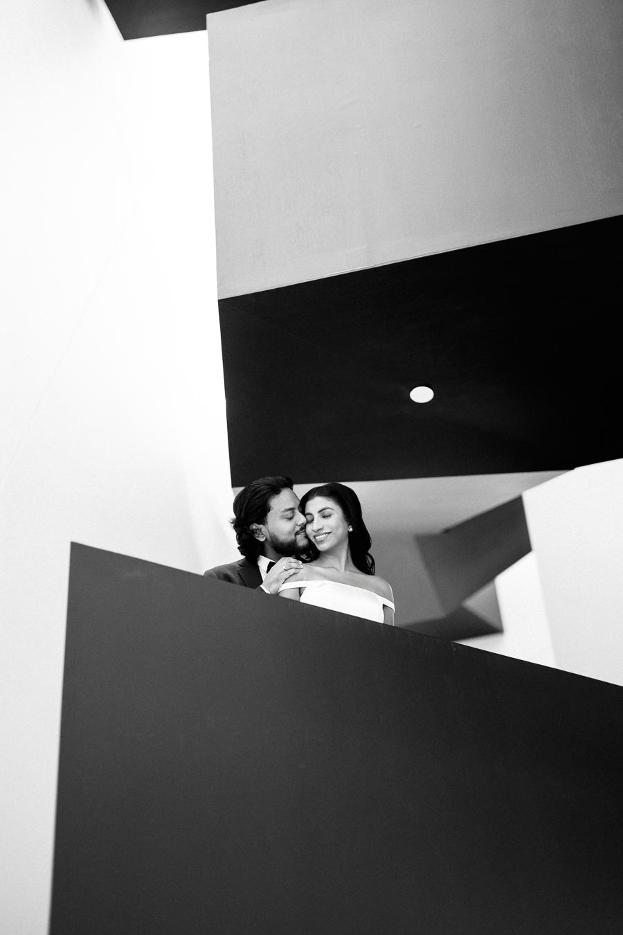 A couple embracing on a modern architectural staircase, captured in a black and white photograph with geometric lines and soft lighting.