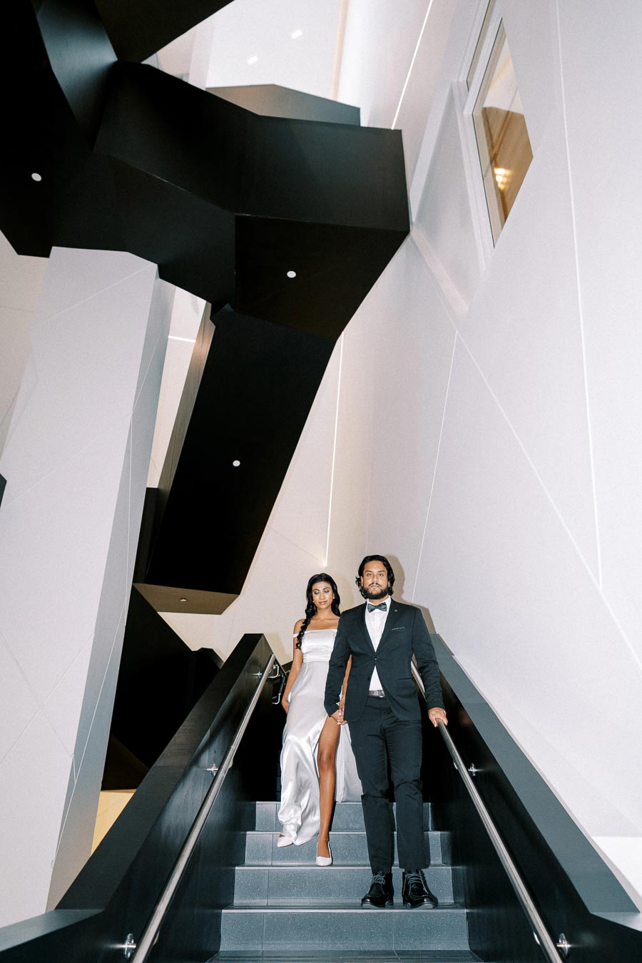 A elegantly dressed couple descends a modern staircase, with geometric black and white architecture enhancing the stylish ambiance.