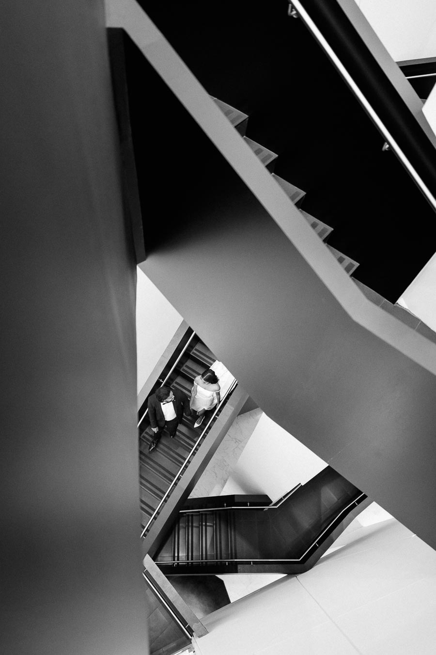 Black and white abstract view of a modern staircase with two people walking, highlighting architectural lines and angles.