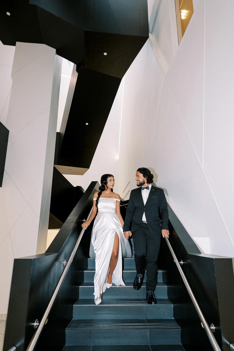 A couple elegantly dressed in formal attire descends a modern staircase with geometric architecture, featuring a woman in a white gown and a man in a black suit, both gazing at each other.