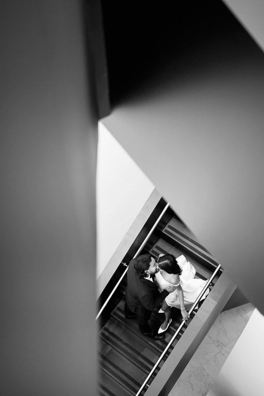 Black and white aerial view of a couple embracing on a staircase, with modern architectural lines creating a dramatic, intimate atmosphere.