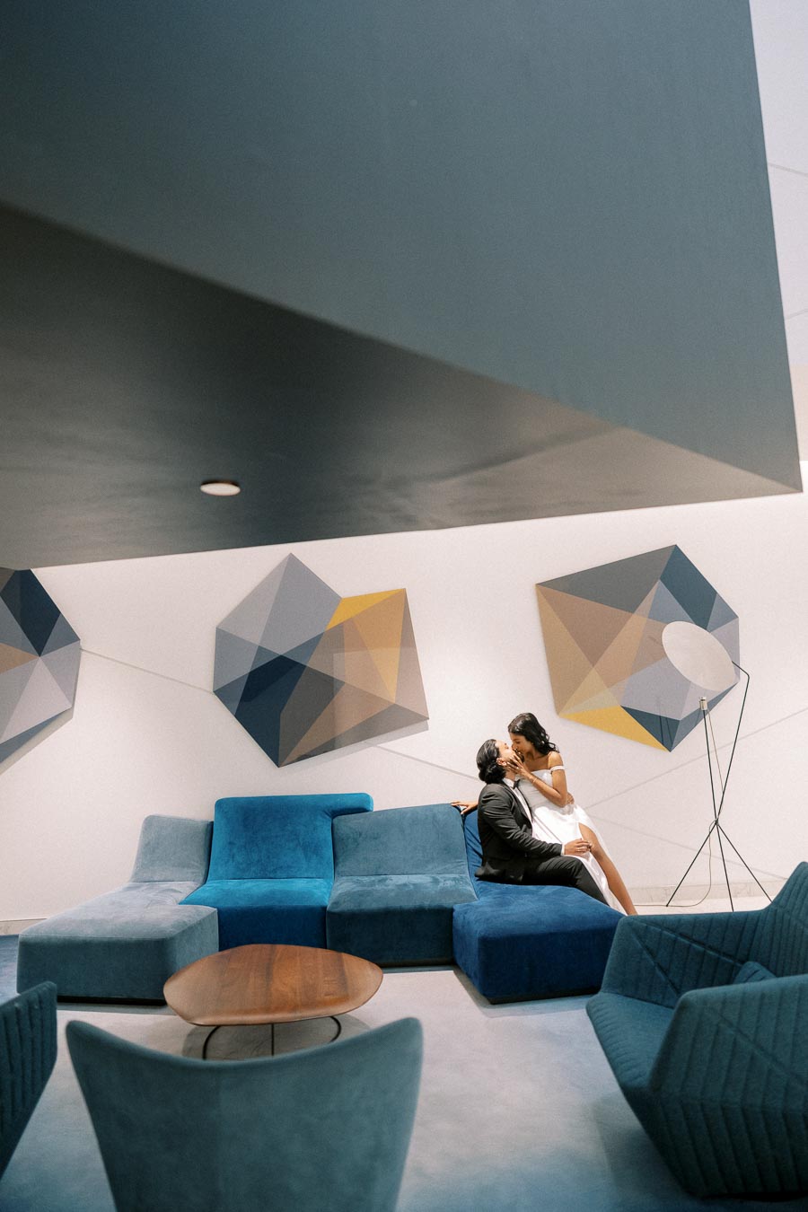 Contemporary lounge area with modern blue sofas and abstract geometric wall art. A couple embraces on the sofa, dressed elegantly, adding a romantic touch to the stylish interior.