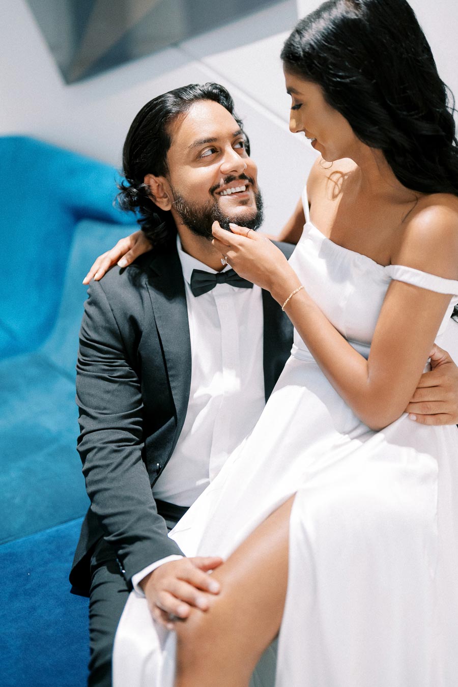 A couple sharing an intimate moment, with the woman in a flowing white dress sitting on a man's lap in a black suit, both smiling at each other on a blue sofa.