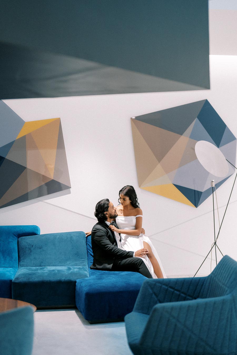 A couple in formal attire sitting on modern blue couches in a contemporary setting with geometric wall art.