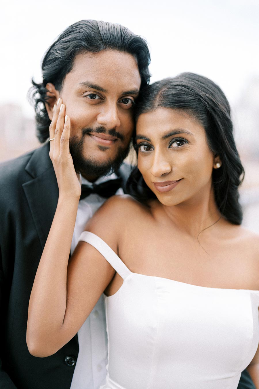 Smiling couple in formal attire with the woman wearing a white off-shoulder dress and the man in a black suit, showcasing a tender moment outdoors.