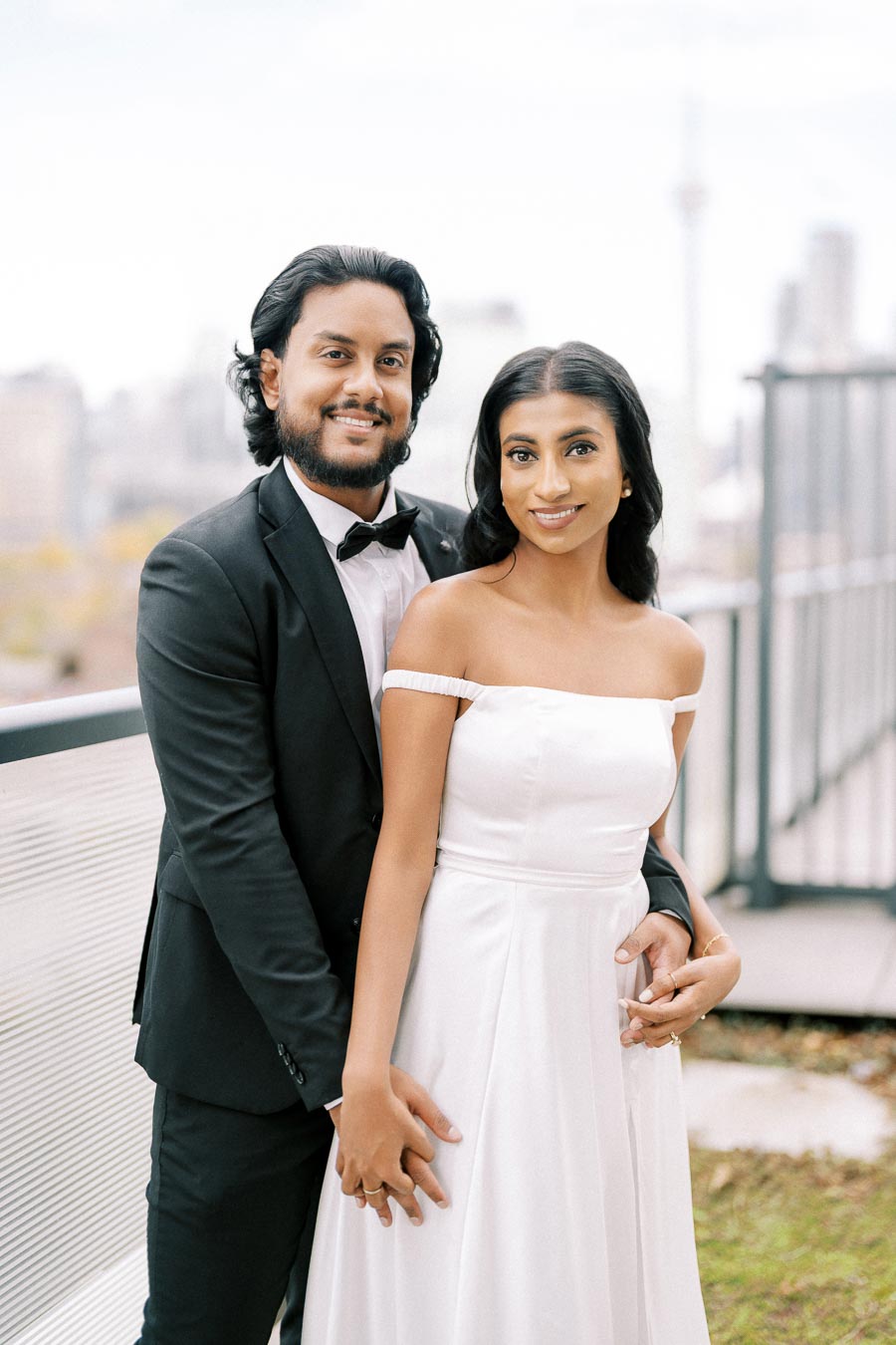 A happy couple posing for a wedding photo on a rooftop with a city skyline in the background. The bride is wearing a white off-shoulder wedding dress, and the groom is dressed in a black tuxedo.