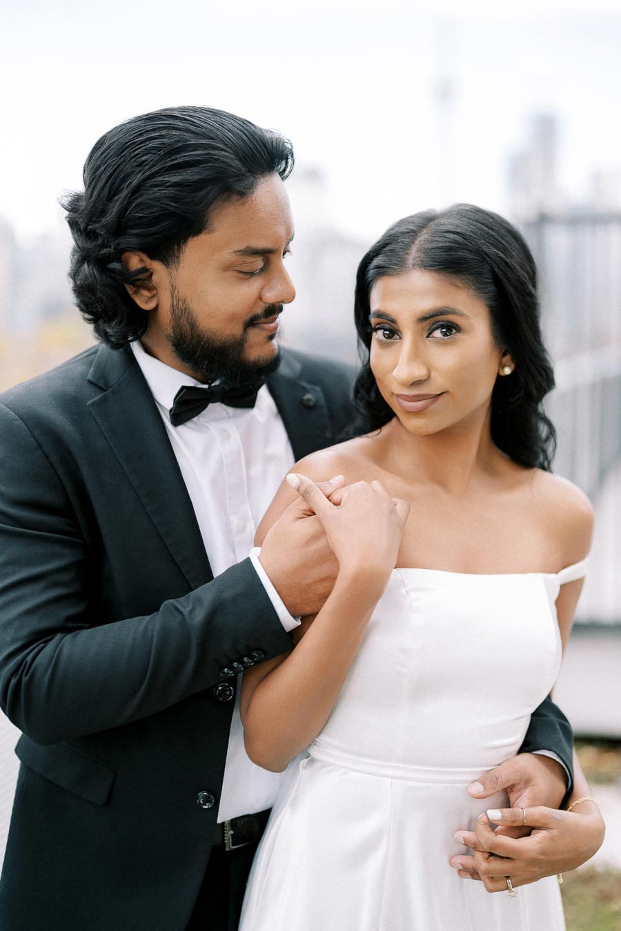 A couple dressed in formal attire, with the man in a black suit and bow tie embracing the woman in a white dress, standing together against a blurred cityscape backdrop.